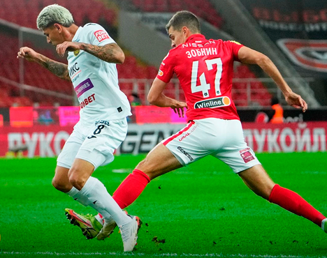 На изображении два футболиста в момент борьбы за мяч на футбольном поле. Один одет в белую форму, второй – в красную. Действие происходит на стадионе, на заднем плане видны трибуны